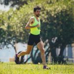 Full length of young bearded sportsman in activewear running on grassy lawn in city park near green trees during outdoor workout
